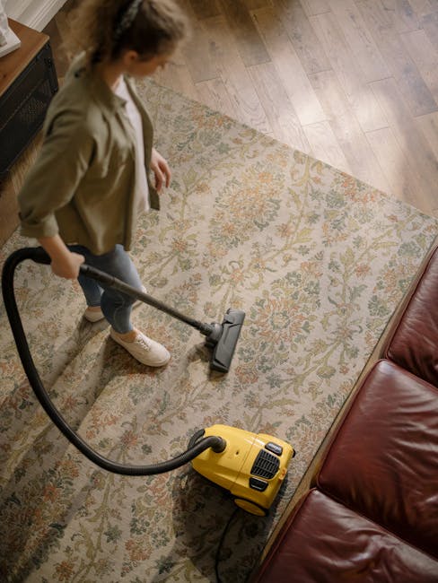 A woman performing surface cleaning on a patterned area rug in a living room. She is using a yellow vacuum cleaner with a hose attachment to deep clean the carpet, which features intricate floral motifs in shades of beige, green, and orange. The room has wooden flooring surrounding the rug, and a red leather sofa is partially visible to the right. The lighting appears natural, highlighting the cleanliness and freshness of the space. The scene exemplifies domestic cleaning with a focus on maintaining hygiene and detailed surface sanitisation, consistent with services offered by Carpet Cleaning Holland Park.