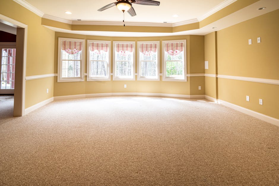 A person is operating a yellow portable carpet cleaner on a patterned area rug in a residential room, with the device's control panel and vent visible. The room features wooden flooring partially visi