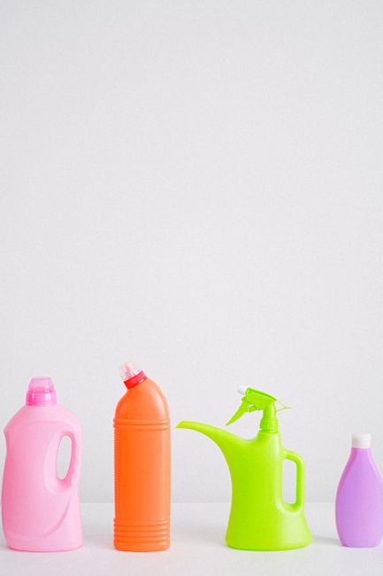 A person wearing a pink rubber glove is holding a pink plastic bottle, likely a cleaning product, against a matching pink background. The bottle is upright, with the hand gripping the handle, and the 