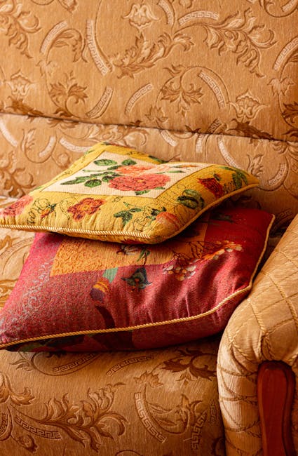 Close-up image of two decorative cushions placed on a fabric-upholstered sofa with a floral pattern. The top cushion features a floral tapestry design with pink, green, and yellow tones, while the bottom cushion showcases a darker, textured fabric with a multicolored design. The sofa's upholstery has a beige or light brown color with a subtle, ornate pattern, and the scene is illuminated by soft, natural lighting. The cushions appear clean and well-maintained, reflecting professional domestic cleaning services provided by Carpet Cleaning Holland Park for upholstery surface cleaning and sanitisation.