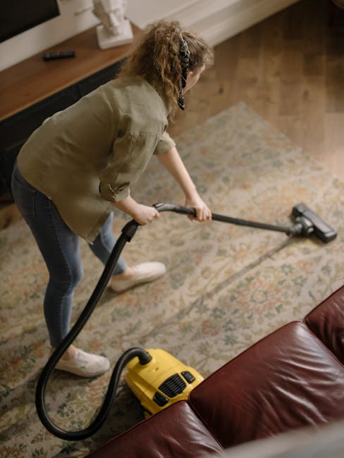 A person kneeling on a patterned carpet in a residential room, operating a yellow portable vacuum cleaner with a black hose. The individual, dressed in a beige jacket and blue jeans, is in the process of deep cleaning the carpet surface, with their hand on the vacuum's controls. The room features warm lighting, a wooden floor visible near the carpet, and the vacuum cleaner's compact design and attachments suggest a focus on surface cleaning and maintenance, which aligns with the upholstery cleaning services offered by Carpet Cleaning Holland Park for Holland Park Estate W11 homes. The scene emphasizes hygiene and professional cleaning practices for domestic spaces.