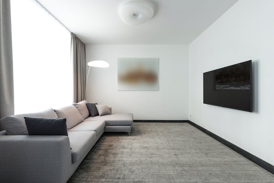 A modern living room with a large, L-shaped grey fabric sofa adorned with multiple light grey cushions. In front of the sofa, there are two white nested coffee tables, one holding a closed dark grey n